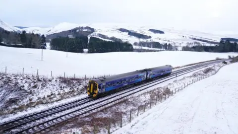 PA A blue train travelling along a snowy track, surrounded by snow-covered fields in Scotland.