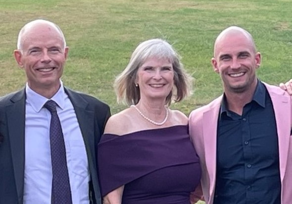 An older man wearing a suit and tie and a woman wearing a purple dress smile as they pose next to a younger man wearing a pink suit and blue shirt. 