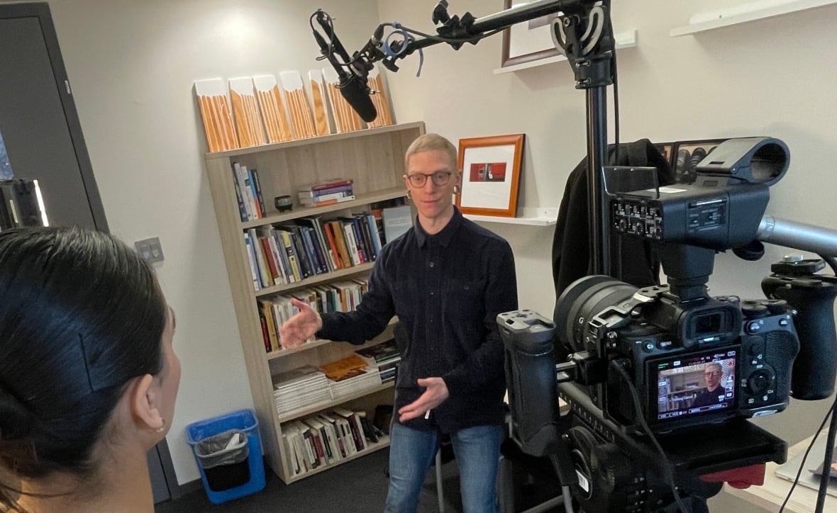 A blonde man with glasses speaks to a young woman in an office while facing a camera.