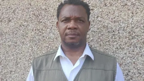 BBC Charles Ehikioya seen standing in front of a textured beige wall. He is wearing a white collared shirt under a green vest with a small brown detail near the chest pocket.