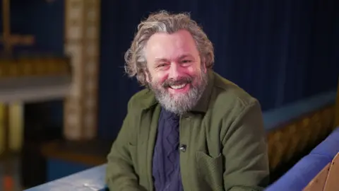 BBC Michael Sheen, who has long grey hair and a beard, smiles to camera while sitting in a theatre 