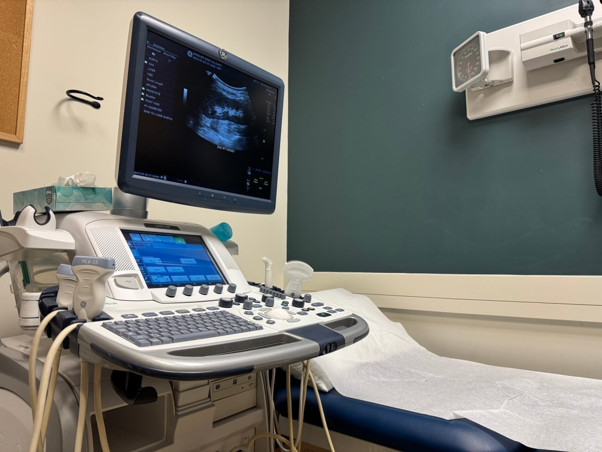 An ultrasound machine next to an empty patient bed. The monitor displays an ultrasound image of an abdomen.