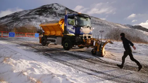 PA Media A snow plough driver at the closed snow gates on the A93 in Spittal of Glenshee - a man is walking across the snow towards the vehicle. 