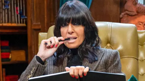 BBC/Studio Lambert Claudia Winkleman on The Traitors, holding a notebook and pen