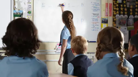 Getty Images A school girl wearing a light blue shirt and grey skirt, with a long blonde plait, fills in a letter on a white board. Her classmates are watching her complete the word puzzle in their classroom. 