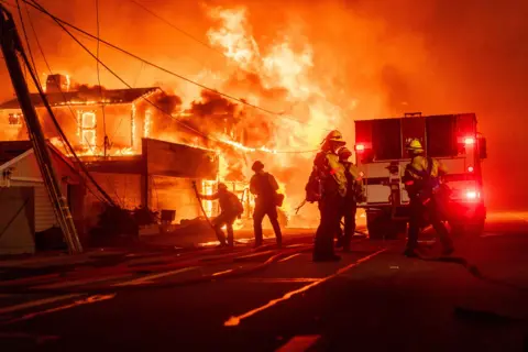 Kyle Grillot/Bloomberg via Getty Images Silhouetted firefighters gather in front of a fire engine next to a burning house in the Pacific Palisades area