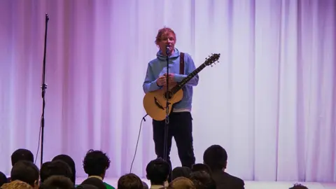 Ed Sheeran Foundation Ed Sheering on stage in front of school pupils. he is holding a guitar and is wearing a lights blue hoodie and dark trousers