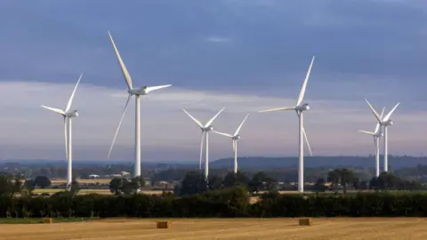 Getty Images Seven large white wind turbines tower over a sunny countryside scene, including a golden harvested wheat or corn field in the foreground.