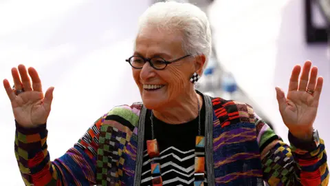 Reuters Rosita Missoni, wearing a Missoni cardigan in a distinctive rainbow pattern with a bold statement necklace and earrings, waves at viewers during a dashion show in 2017