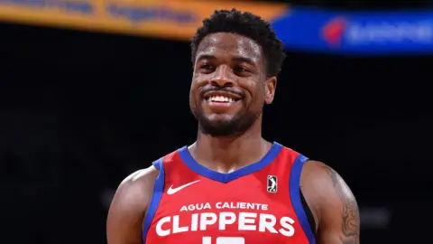 Getty Images Malik Dunbar smiles while on court in a basketball game. He is wearing a red sports kit with Agua Caliente Clippers written on it in white typeface.