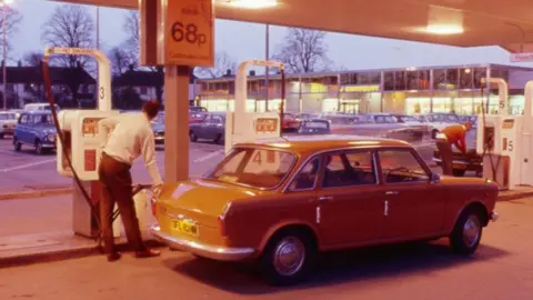 The Sainsbury Archive A man filling a car at a petrol station in 1975. He has his back to the viewer, is facing a white petrol pump and is holding the petrol feed which is filling a an orange car. He is wearing a white shirt tucked into brown trousers. Above his car is a sign saying petrol 68p