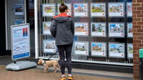 Getty Images Woman in running gear and a coat, with a small dog on a lead, looks at adverts in an estate agents window.