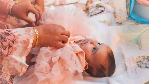 Family handout Sophia Kelemen lies on a changing mat. She is wearing a peach coloured outfit with ruffles and has a dummy in her mouth. A woman's hands can be seen adjusting her outfit.