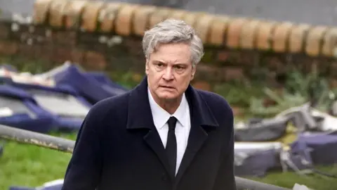 PA Media Colin Firth walking with a concerned expression. Wreckage can be seen in the background, and he is wearing a black coat and tie, with a white shirt.