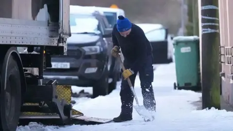 PA Media A man wearing a cap and heavy clothing uses a shovel to clear snow from a pavement in a street where cars and a lorry are parked.