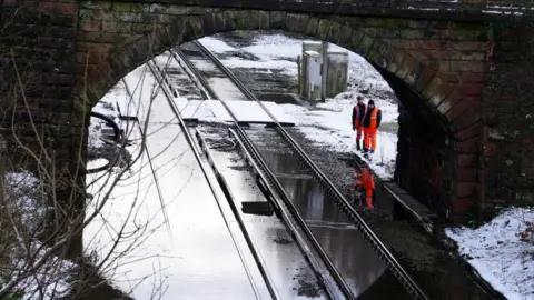 PA Two workmen in orange high-vis gear survey a flooded railway track passing under a bridge, surrounded by snow.