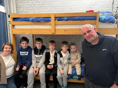 BBC Mother Sam stands to the far left wearing a white T-shirt, cardigan and jeans. She has a short bob hairstyle. Next to her, sitting on the bottom bunk bed are her five sons all lined up together wearing tracksuits. Their father Jason is standing on the right wearing a navy hooded top. He has short grey hair.