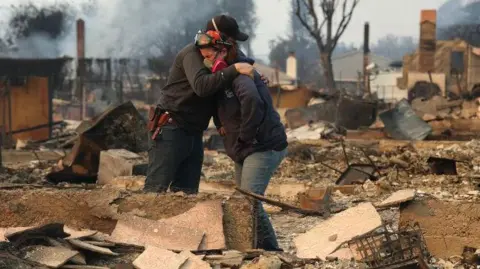 Getty Images Two people embrace as they inspect a family member's property that was destroyed by the Los Angeles wildfires.