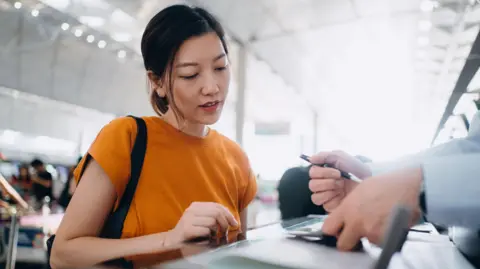 Getty Images Young woman wearing an orange T-shirt checking in at an airline check-in counter at an airport