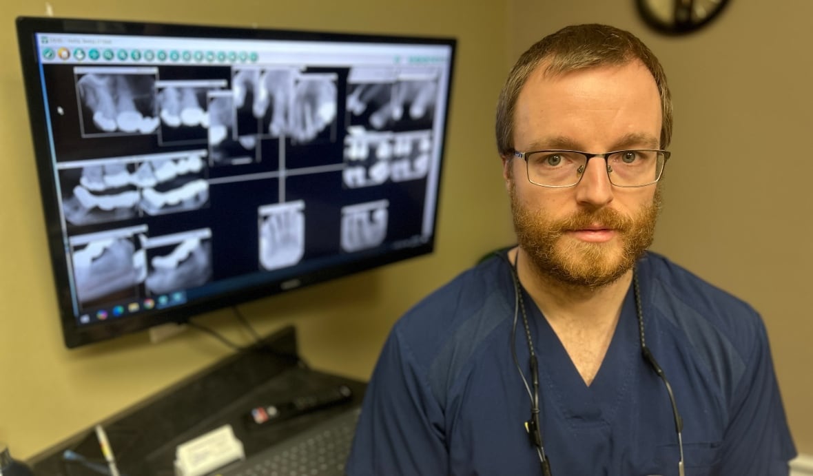 Dr. Brandon Doucet wears scrubs and sits infront of a tooth x-ray at a dental office in Amherst, Nova Scotia.