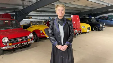 BBC A woman with short blonde hair stands in front of a row of classic cars of a variety of colours. She is wearing a long black leather coat.